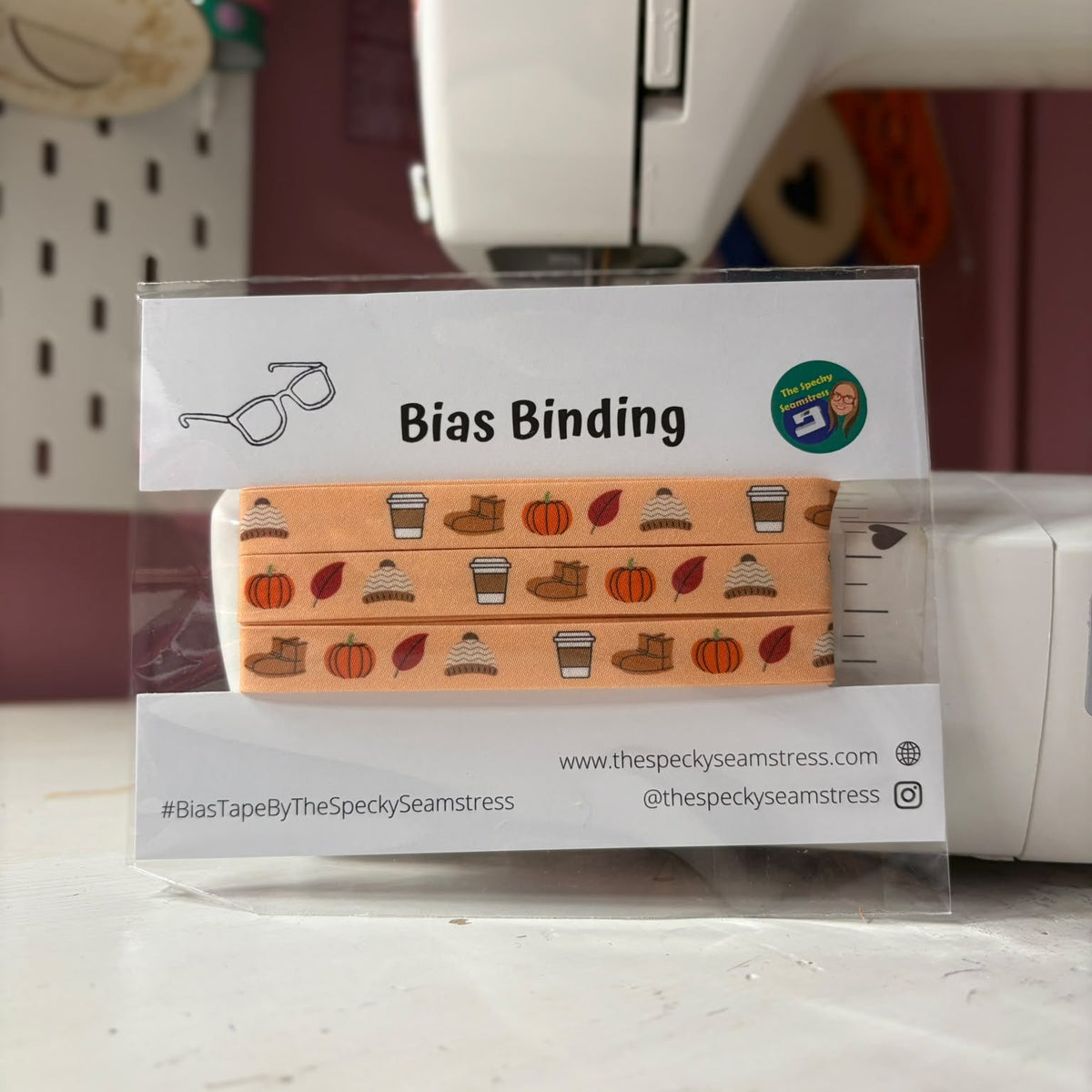 A packaged bias binding tape roll with a fall-themed pattern of pumpkins, leaves, and boots, displayed in a clear plastic sleeve on a sewing machine table.