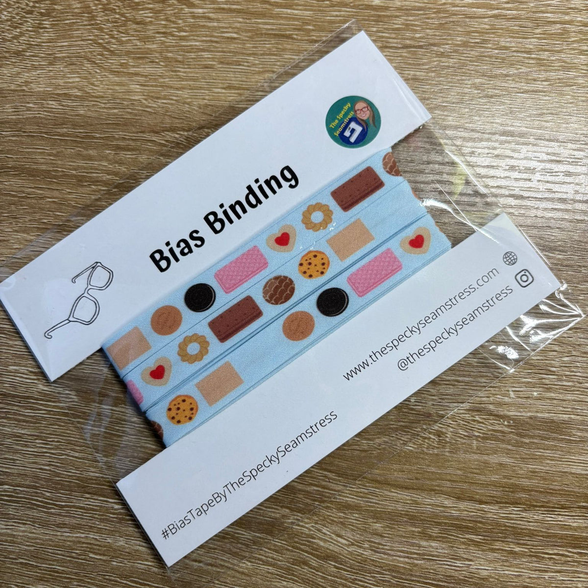 Light blue bias binding with cartoon biscuits on a white backing card in a clear bag, on a wooden background.