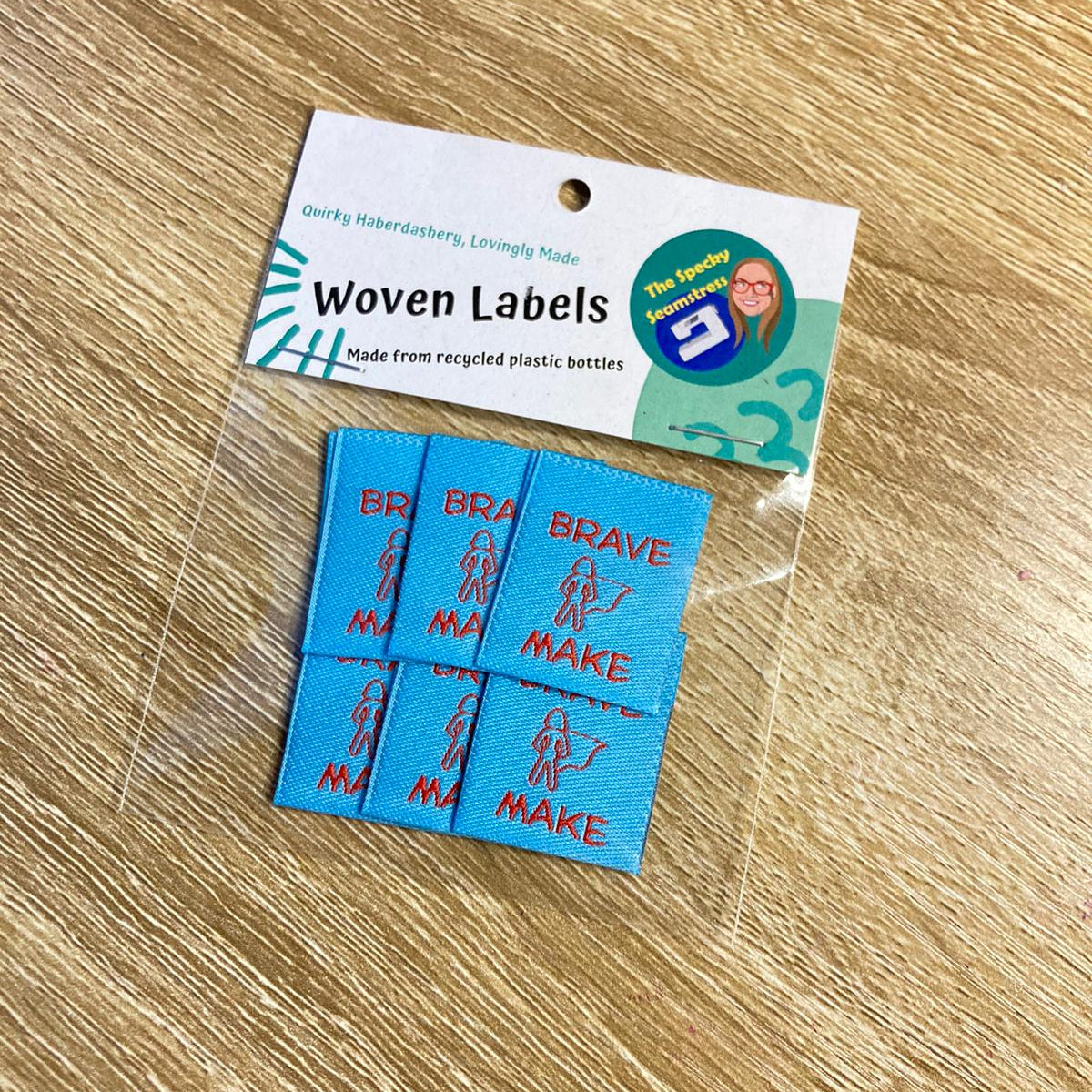 Set of 6 blue woven 'Brave Make' labels with a packaging card on a wooden surface.