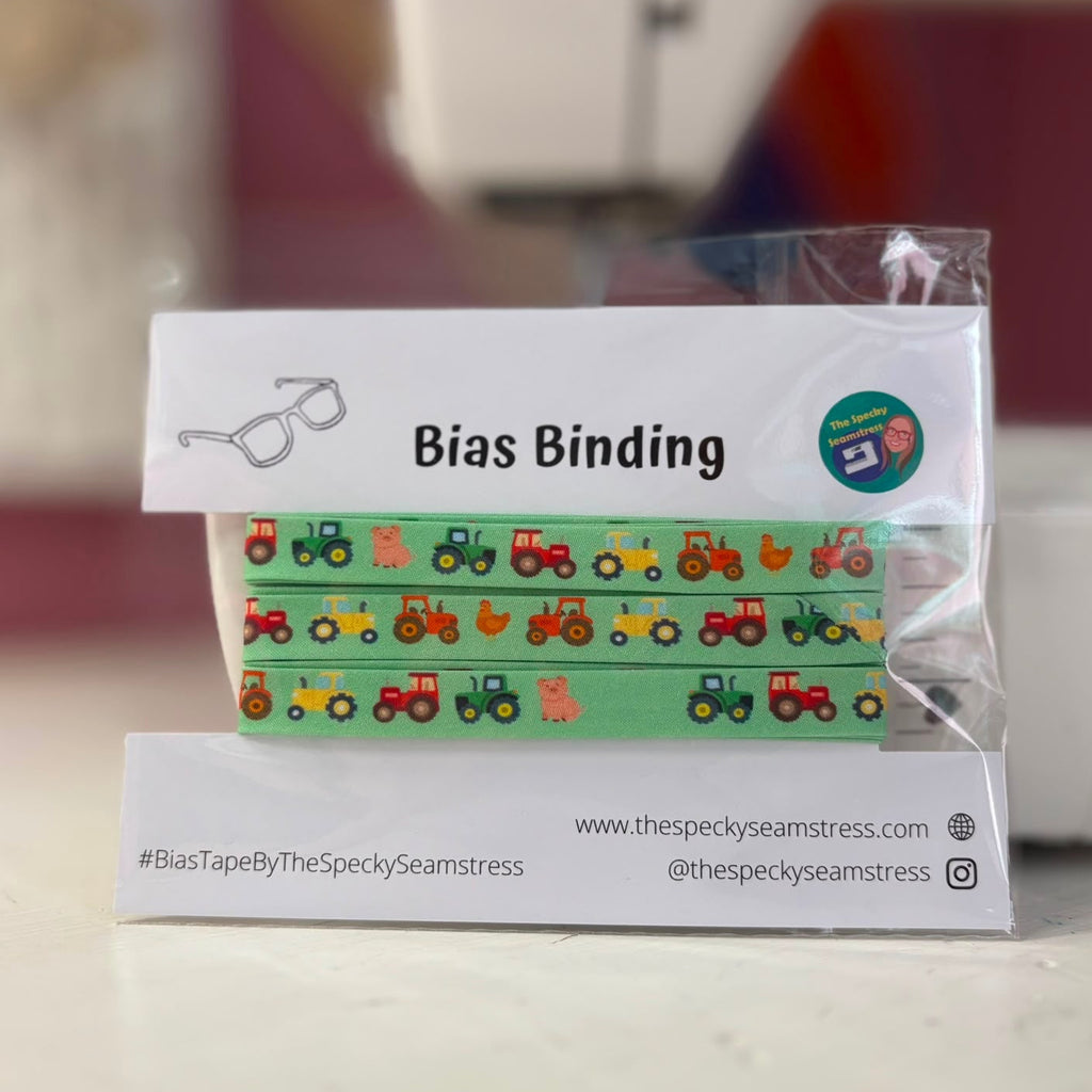 A green bias tape featuring tractors, pigs and chickens in a repeating sequence, on a white backing card, in front of a sewing machine. 