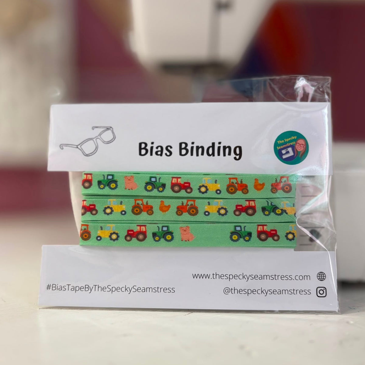 A green bias tape featuring tractors, pigs and chickens in a repeating sequence, on a white backing card, in front of a sewing machine. 