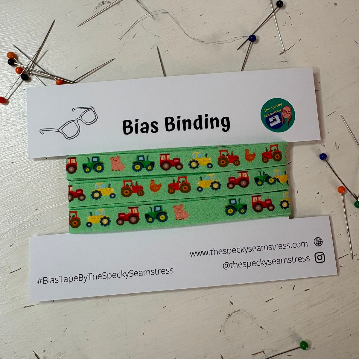 A green bias tape featuring tractors, pigs and chickens in a repeating sequence, on a white backing card, in front of a sewing machine. 