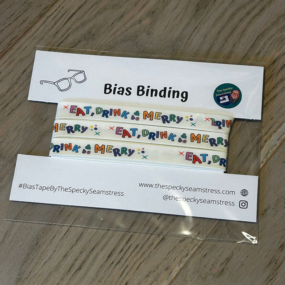Eat, Drink and Be Merry Bias binding tape with colorful text design on a wooden surface.