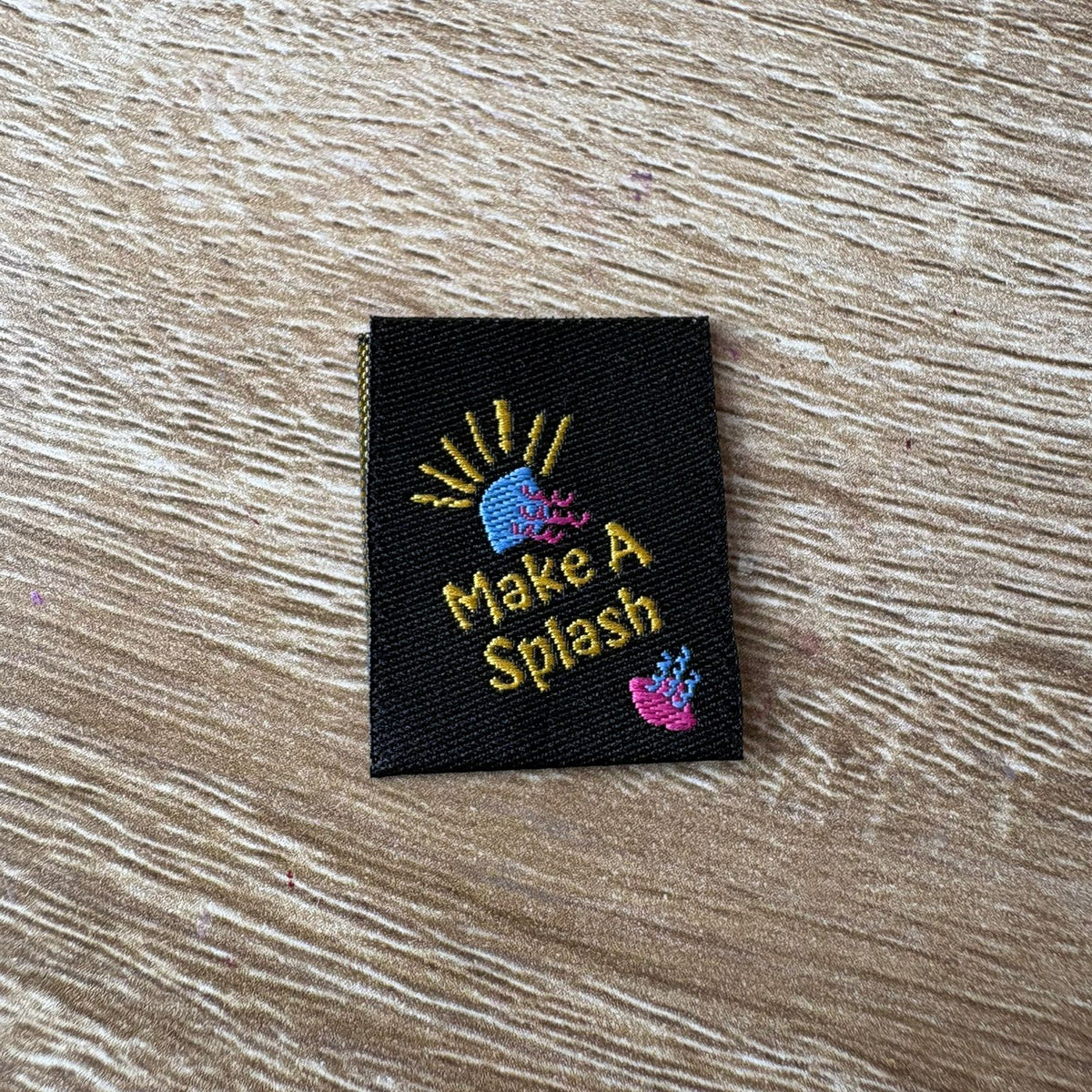 Embroidered label with 'Make A Splash' text on a wooden surface