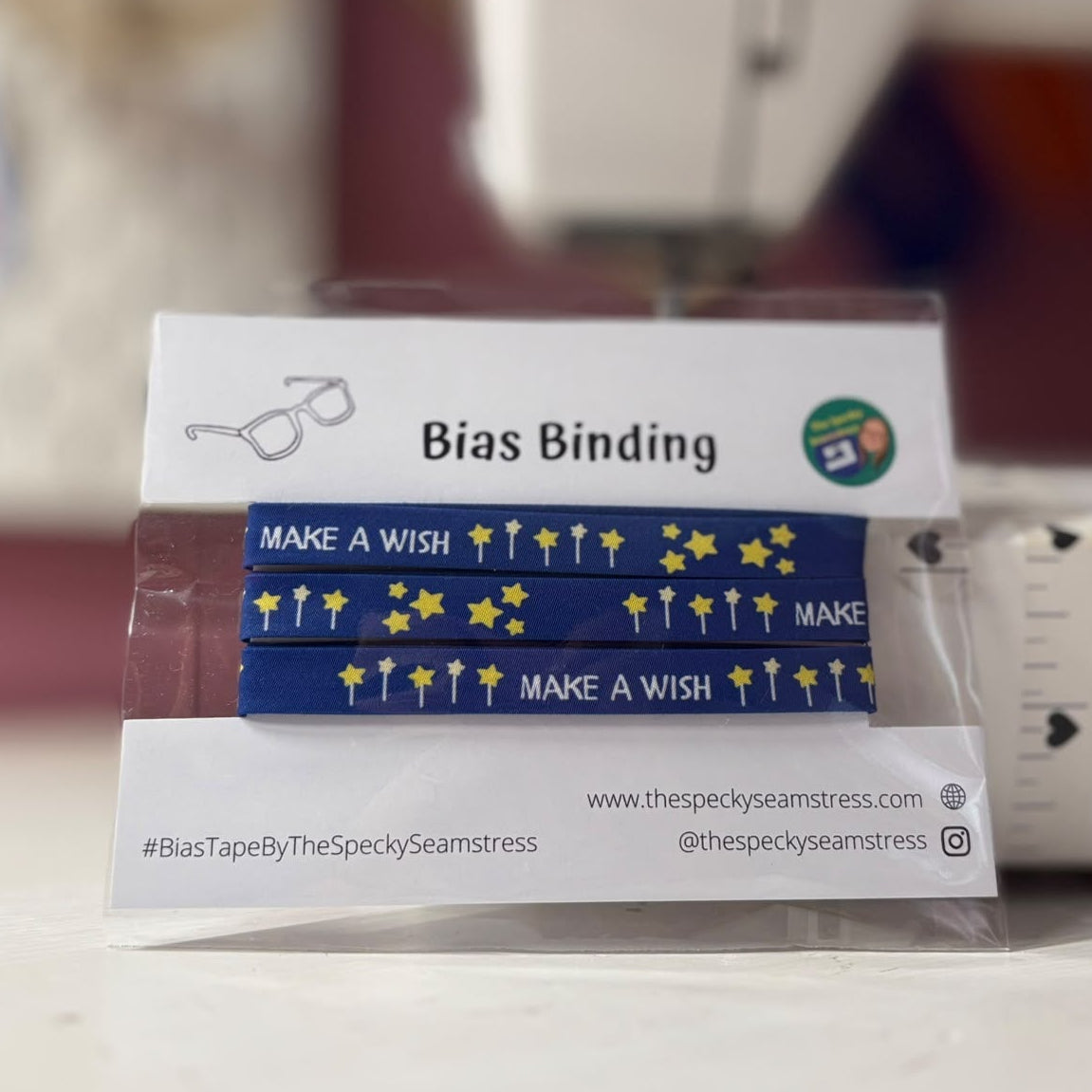 A package of bias tape ribbons with the text 'MAKE A WISH' in yellow on a blue background, labelled 'Bias Binding' on the card, with a sewing machine blurred in the background.