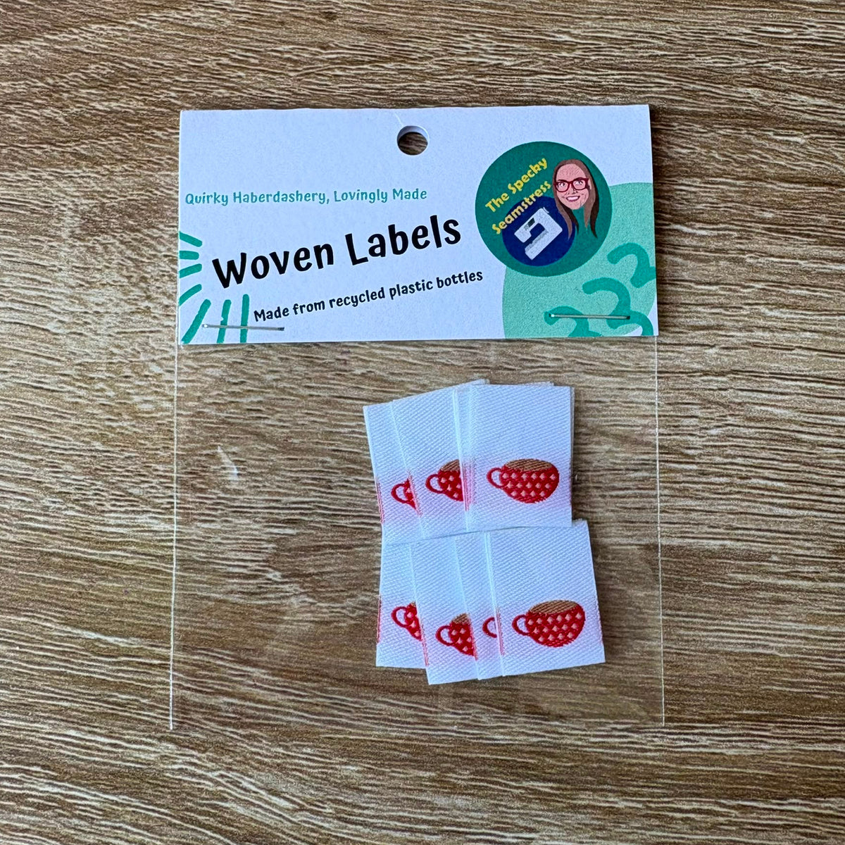 Woven labels with red and white teacup design in a clear packet with a card top, placed on a wooden surface.