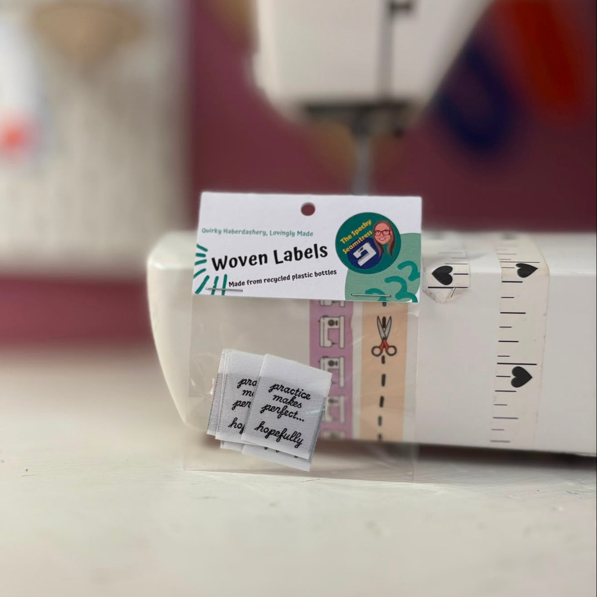 A small clear plastic package labeled 'Woven Labels' containing tiny handwritten fabric labels, photographed in front of a sewing machine on a tabletop.