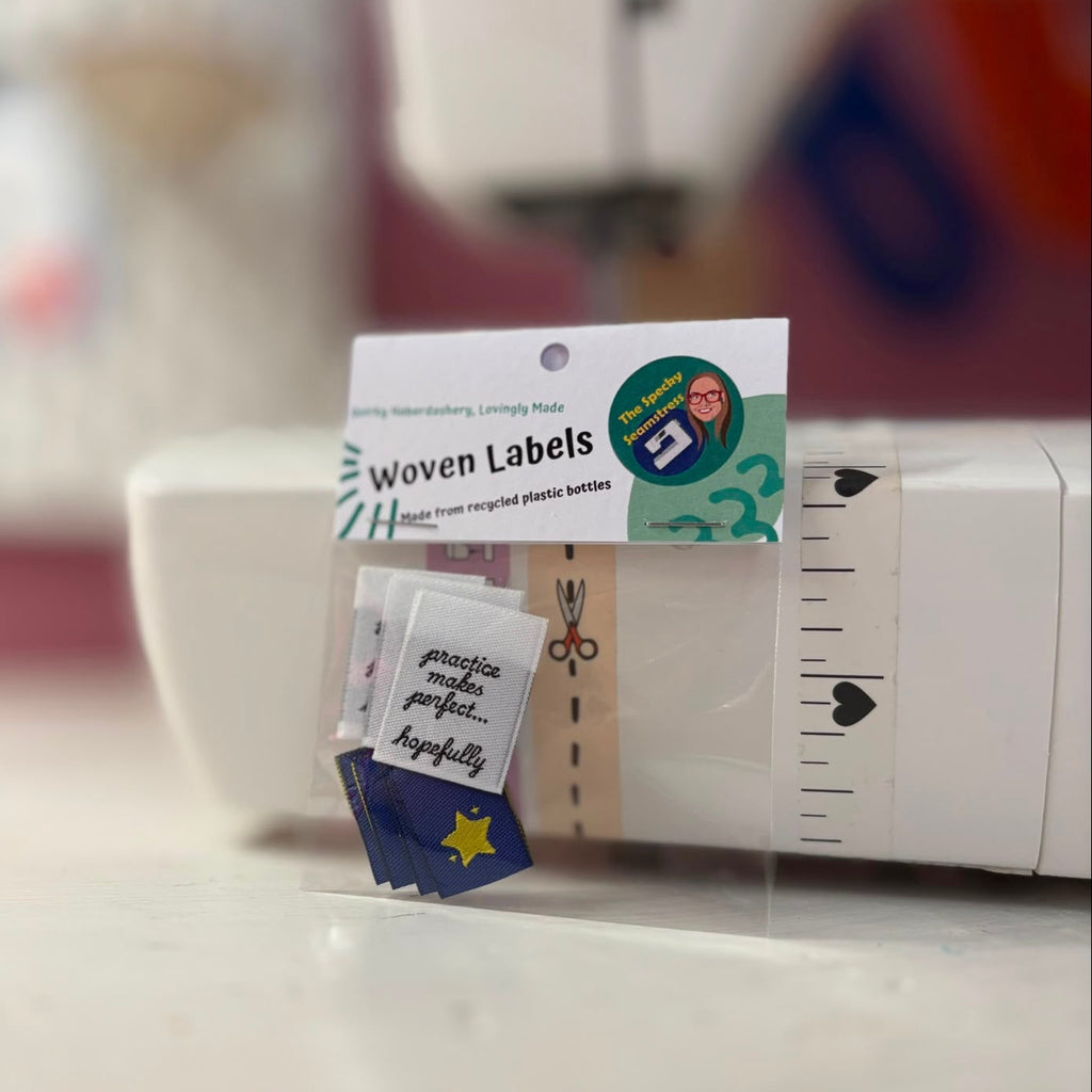 A small clear plastic bag labeled 'Woven Labels' containing several small fabric labels, placed in front of a white sewing machine. Some labels are blue with a yellow star and some are white with black script.