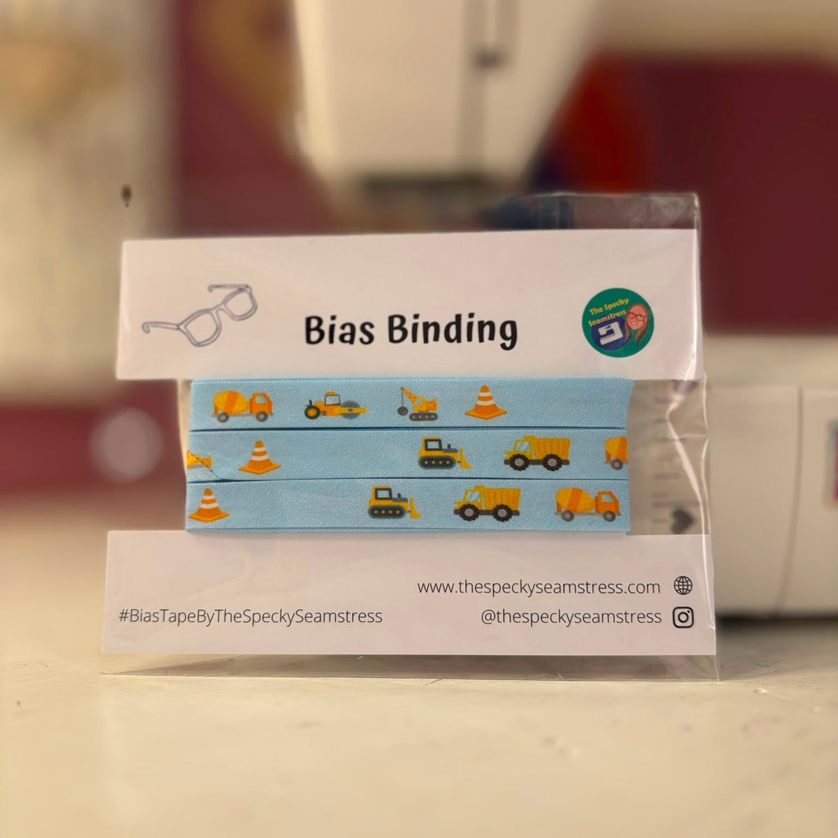 A blue bias tape with yellow construction vehicles and traffic cones, on a white cardboard backing, in front of a sewing machine.